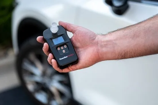 Person holds ignition interlock device in front of white car.